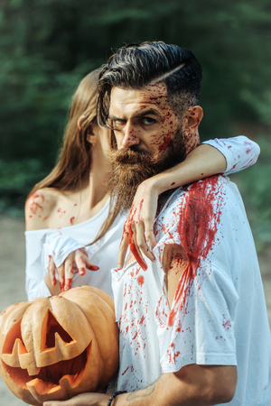 young halloween couple of bearded man with beard and sexy pretty girl with red blood and traditional autumn holiday symbol of orange spooky pumpkin outdoor in deep green forest or woodの写真素材