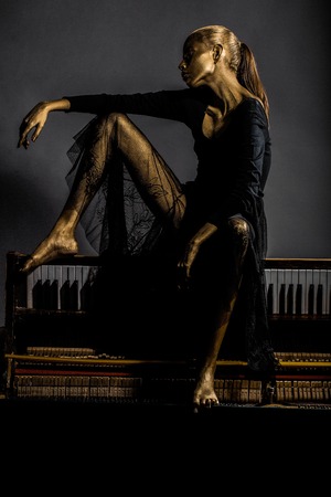 sexy golden slim woman or girl has pretty face with makeup and body art metallized color in black dress sitting on old wooden piano with keyboard on grey backgroundの写真素材
