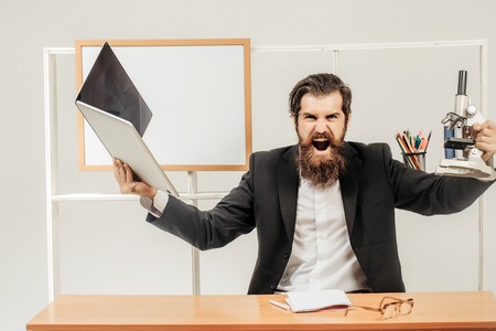 Bearded scientist in suit shouts with laptop and microscope in his hands at desk with exercise book pen and glassesの写真素材