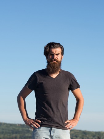 Handsome man hipster with brunette hair long beard and moustache in casual tshirt stays outdoors on sunny day on blue skyの写真素材
