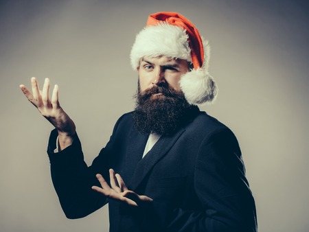 Handsome young man with long beard and moustache in red santa claus hat and jacket with raised hand in studio on grey backgroundの写真素材