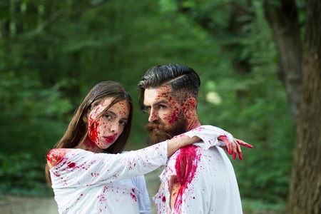 young halloween couple of bearded man with beard and sexy pretty girl with red blood as traditional autumn holiday symbol outdoor in deep green forest or woodの写真素材