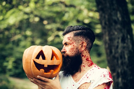 young halloween bearded man with beard and red blood with traditional autumn holiday symbol of orange spooky pumpkin outdoor in deep green forest or woodの写真素材