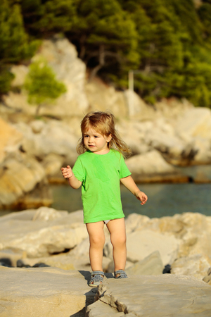 Cute baby boy child with curly blond hair in green shirt plays at rocky sea beach on summer dayの写真素材