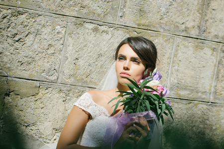 young sexy woman or girl bride with makeup on pretty face and veil in hair holding wedding bouquet or posy of eustoma and rose flowers sunny outdoor on stony wall backgroundの写真素材