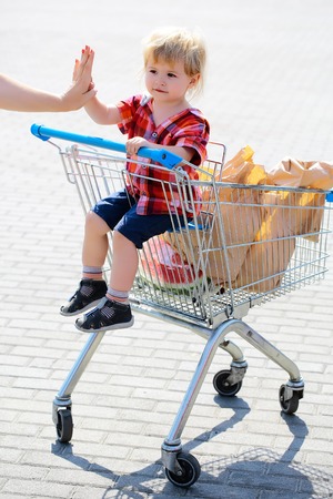 Cute baby boy blond child in plaid shirt sits in shopping trolley and eats cookie from paper packet outdoors on sunny dayの写真素材