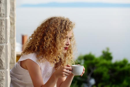 pretty woman with curly blond hair stands on balcony with coffee cup summer day on natural backgroundの写真素材
