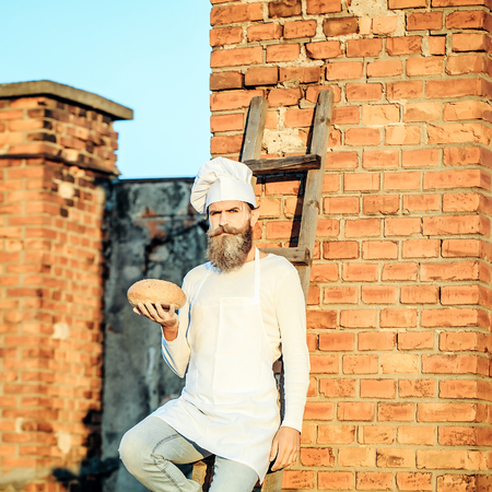 Handsome bearded man cook chef in white uniform with long beard holding bread in hands on sunny day...
