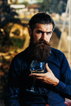handsome bearded man with stylish hair mustache and beard on serious face holding glass of white wine on city backgroundの写真素材