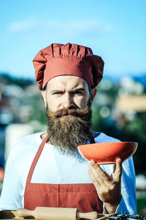 Handsome bearded man cook chef uniform and red hat with long beard standing near table with rolling pin on it and holding bowl in sunny day outdoor on blue sky backgroundの写真素材