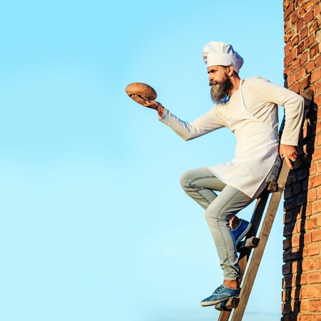 Handsome bearded man cook chef in white uniform with long beard holding bread in hands on ladder on sunny day outdoor on blue sky background, copy spaceの写真素材