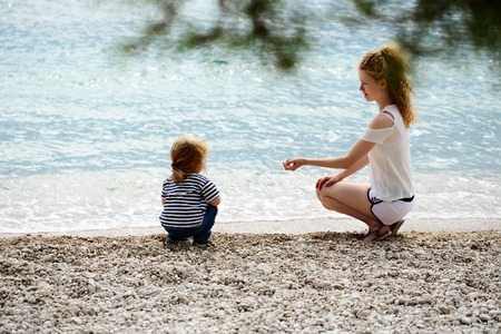 Mother young woman and son baby boy sit on pebble beach near blue sea windy weather on natural backgroundの写真素材