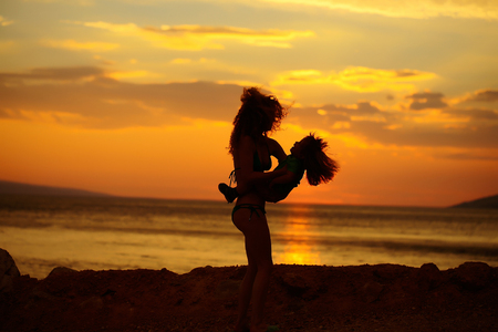 Mother young woman and son baby boy silhouettes plays on beach near sea or ocean water in evening or twilight sunset summer day on natural backgroundの写真素材