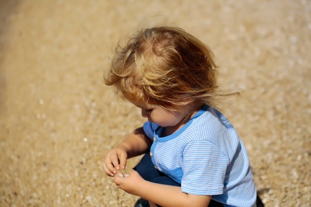 Cute baby boy child with curly blond hair in blue shirt sits on haunches and plays with sand on...