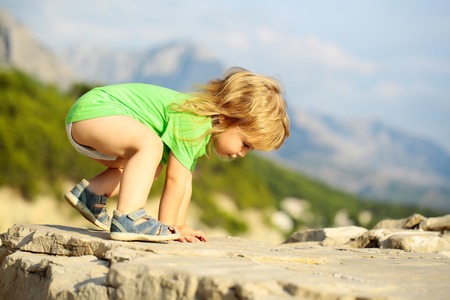 Cute baby boy child with curly blond hair in green shirt plays on rocks on sunny summer day on natural backgroundの写真素材