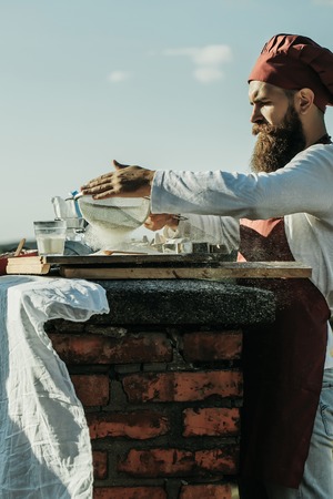 Handsome bearded man cook chef in uniform and red hat with long beard cooking in sunny day outdoor on blue sky backgroundの写真素材