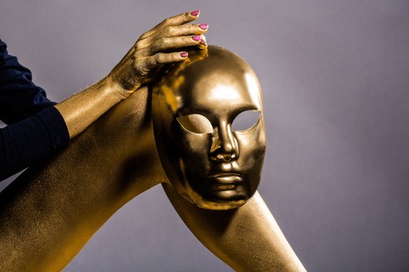 sexy golden female leg and hand with makeup or body art metallized color holding masquerade or carnival mask on grey background, copy spaceの写真素材