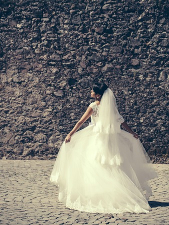 pretty bride woman in elegant white lace dress and veil with wedding bouquet on masonry wallの写真素材