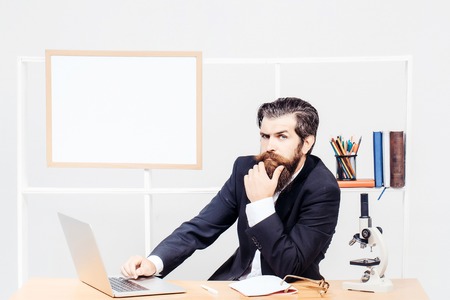 Bearded researcher in black classic suit typing on computer sitting at desk with microscope at workplaceの写真素材