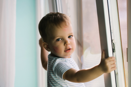 Adorable baby blond boy with red kiss trace on cheek holds window frame and stays on sillの写真素材