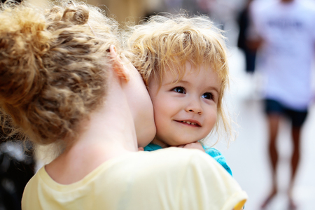 Mother young woman holds happy son baby boy with blonde hair in blue shirt sunny day outdoor in street on blurred backgroundの写真素材