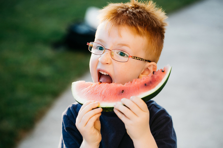 Happy baby boy with red hair in blue shirt and eyeglasses on happy smiling face eating watermelon summer day outdoorの写真素材