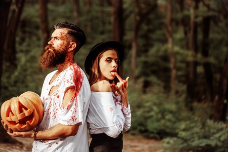 young halloween couple of bearded man with beard and sexy pretty girl in witch hat with red blood and traditional autumn holiday symbol of orange spooky pumpkin outdoor in deep green forest or woodの写真素材