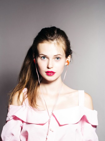 young sexy fashionable woman or girl with long brunette hair and red lips on pretty face in stylish pink dress in studio on grey background with headset on musical playerの写真素材