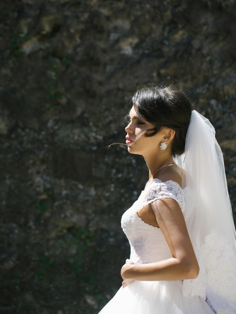 pretty bride woman in elegant white lace dress and wedding veil on stony wallの写真素材