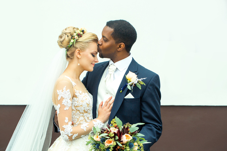 Young man elegant african American groom kisses tenderly beautiful woman happy bride in white dress and veil married couple on wedding dayの写真素材
