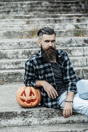 young halloween bearded man with beard and mustache in jeans and stylish checkered shirt sitting on stony stairs with traditional autumn holiday symbol of orange spooky pumpkin outdoorの写真素材