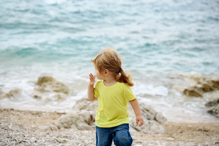 Cute baby boy child walks on pebble beach of stone shore on summer day on blue sea