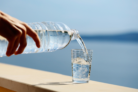 Female hand pours pure drinking water from plastic bottle into glass outdoors on summer day on blue seaの写真素材