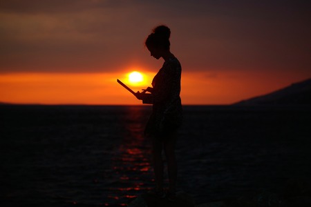 pretty sexy female silhouette of woman or girl with computer or laptop outdoor over dramatic dark sky with clouds and sea or ocean water horizon on evening or twilight on natural sunset backgroundの写真素材