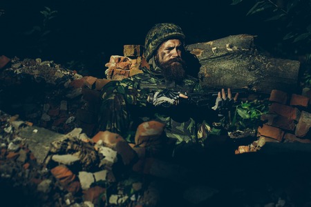 Young soldier with sad bearded face in military helmet and camouflage with gun on ruined bricks background in forestの写真素材