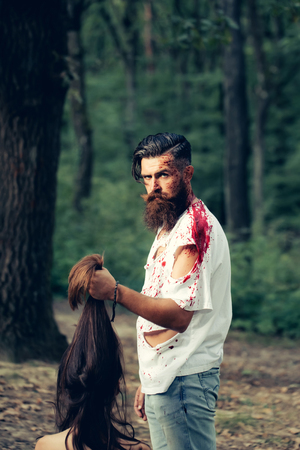 young halloween couple of bearded man with beard and sexy pretty girl with long hair and red blood as traditional autumn holiday symbol outdoor in deep green forest or woodの写真素材