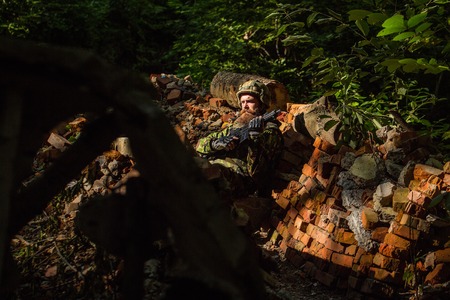 Young soldier with sad bearded face in military helmet and camouflage with gun on ruined bricks background in forestの写真素材