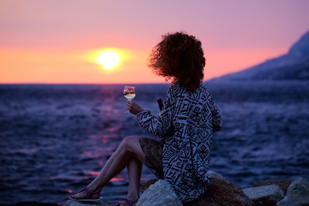 pretty sexy woman or girl with wine glass sits on stone outdoor over twilight sky on beach with sea or ocean water on evening sunset natural backgroundの写真素材