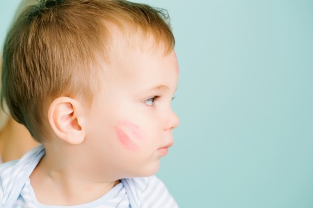 Cute baby blond boy kid child with serious look and red kiss trace on cheek sits on blue wall, copy spaceの写真素材