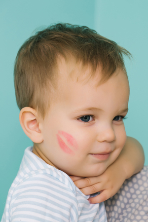 Adorable baby blond boy kid child with red kiss trace on cheek and cute smile on blue wallの写真素材