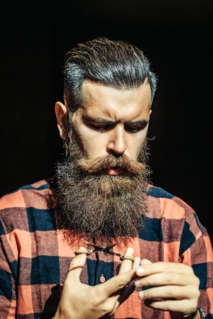 Young man hipster with long beard and moustache handsome face looking at scissors in his hands on black backgroundの写真素材