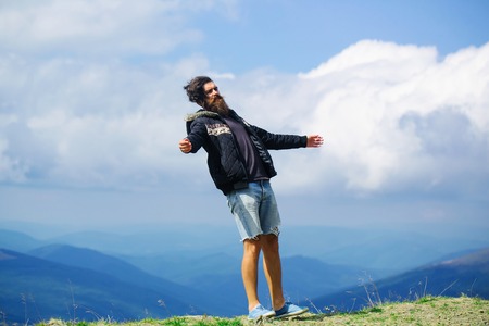 Handsome man hipster with beard and moustache in warm coat and denim shorts stands on mountain top on cloudy skyの写真素材