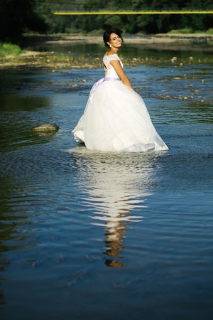 pretty bride pretty woman in long elegant white lace wedding dress stands in lake water sunny day outdoor on natural backgroundの写真素材