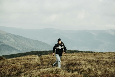 Handsome man hipster with beard and moustache in warm coat and hat runs on mountain top on sunny summer day on cloudy skyの写真素材
