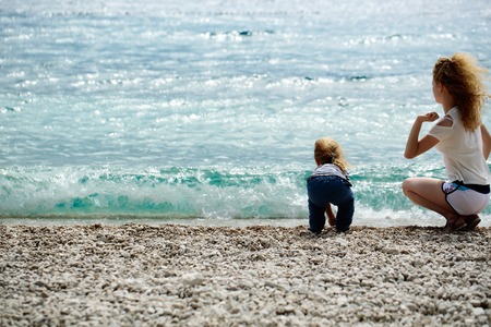 Mother young woman and son baby boy sit on pebble beach near blue sea windy weather on natural background, copy spaceの写真素材