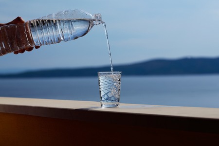 Female hand pours pure drinking water from plastic bottle into glass outdoors on summer day on blue seaの写真素材