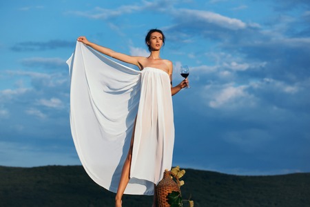Pretty young girl or woman in white sexy dress denuded with red wine in glass wicker bottle vine and grapes on nature over blue skyの写真素材