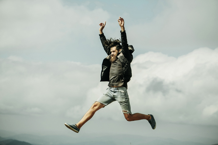 Handsome man hipster with beard and moustache in warm coat and denim shorts jumps on mountain top on cloudy skyの写真素材