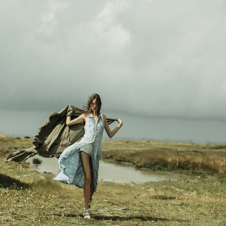 Pretty girl slim sexy female model with long brunette hair in blue dress with blanket walks on summer field on cloudy skyの写真素材
