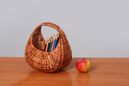 Wicker basket with colorful colored pencils for painting and red ripe apple on wooden brown school desk on grey background studioの写真素材